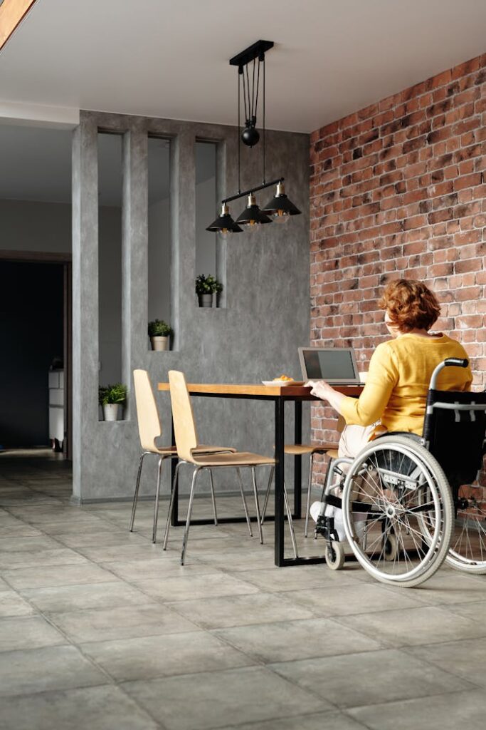 A woman in a wheelchair using a laptop at a stylish home office setup with a brick wall.